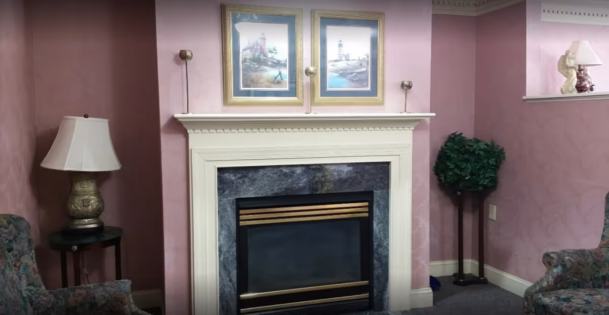 A cozy living room corner featuring a fireplace with a white mantel and marble surround. Above the mantel are two framed paintings depicting coastal scenes. On either side of the fireplace are wall-mounted candle holders. To the left is a table lamp on a small round table next to a floral upholstered armchair. To the right is a tall artificial plant and part of another floral upholstered chair. The walls are painted in a soft pink color with a subtle pattern.