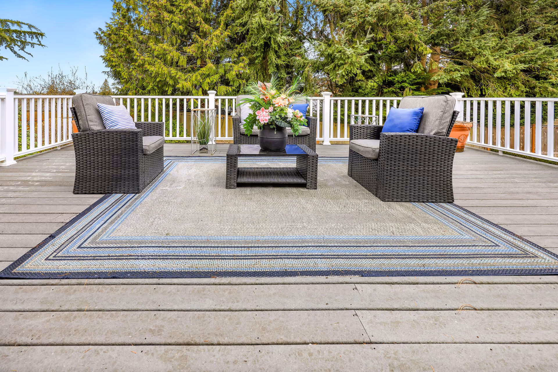 Outdoor deck area with gray wicker patio furniture including two armchairs and a loveseat, all with gray cushions and blue accent pillows. A rectangular glass-top coffee table with a flower arrangement sits on a large outdoor rug with blue and gray stripes. The deck is surrounded by white railing and tall green trees in the background.