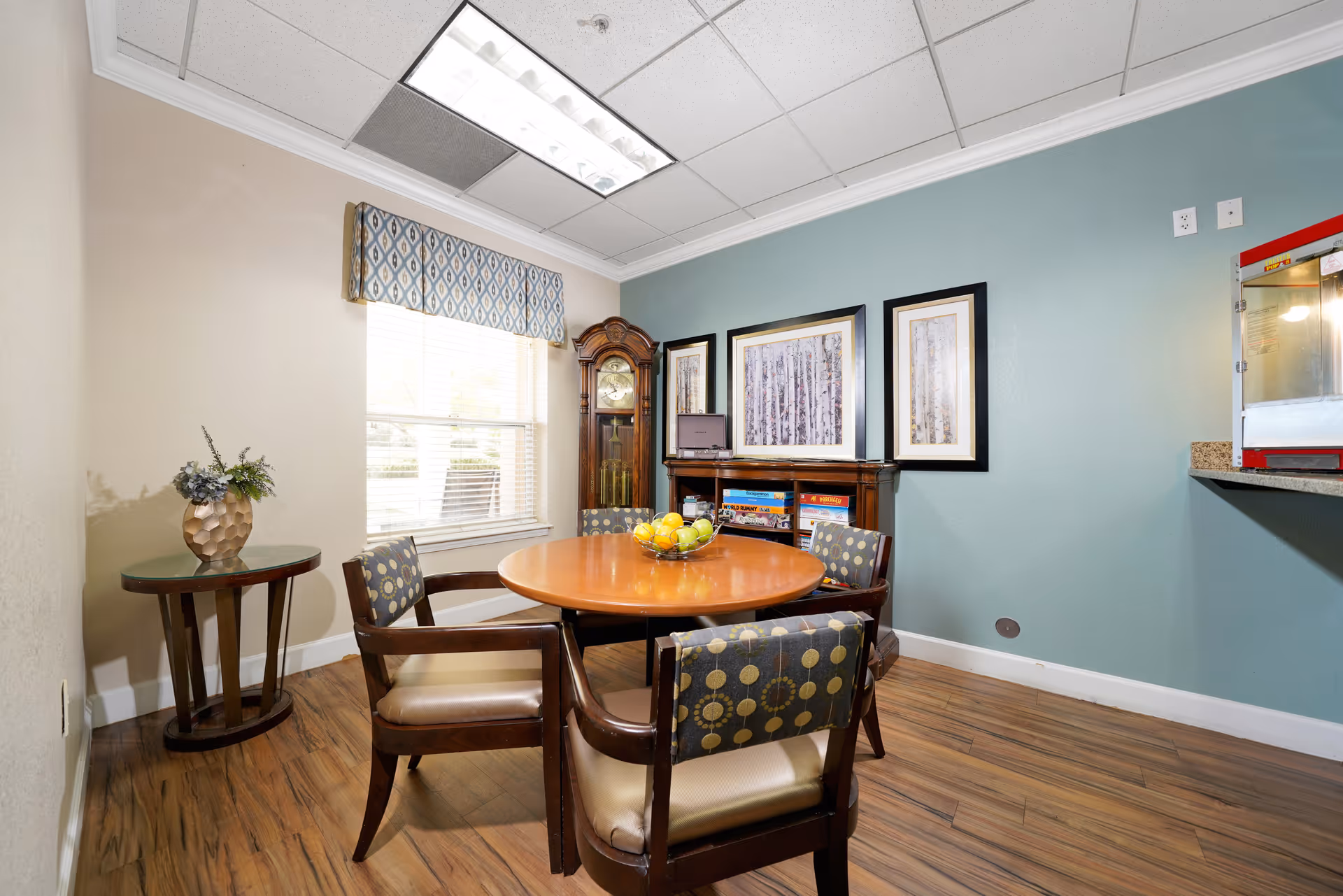 A small dining area with a round wooden table surrounded by four cushioned chairs with patterned backs. A bowl of fruit is placed on the table. Behind the table is a wooden cabinet with board games and a laptop on top, flanked by three framed pictures of birch trees on a blue wall. To the left, there is a window with blinds and a patterned valance, and a small round side table with a decorative vase holding flowers. The floor is wood, and there is a popcorn machine on a counter to the right.
