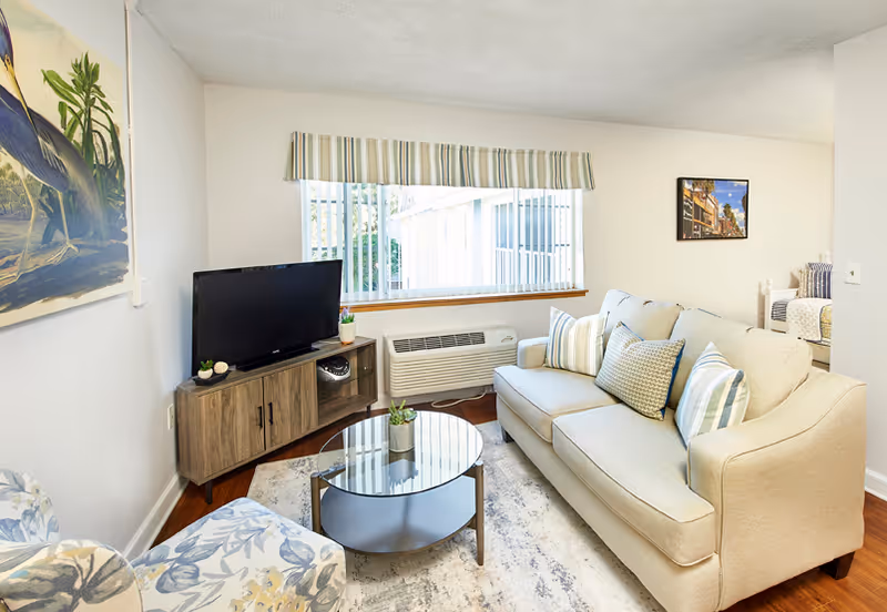 A cozy living room with a beige sofa adorned with striped and patterned pillows, a round glass coffee table with a small plant, a wooden TV stand with a flat-screen television, and a large window with vertical blinds and a striped valance. There is a floral-patterned armchair partially visible and artwork on the walls.