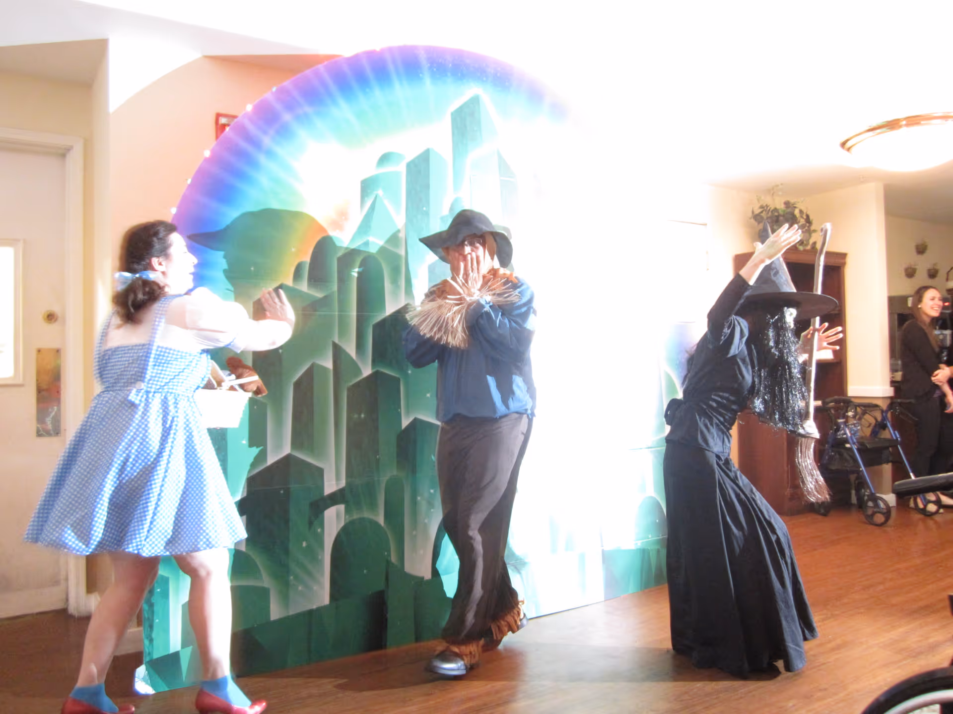 Three people dressed in costumes from The Wizard of Oz, including Dorothy, the Scarecrow, and the Wicked Witch of the West, performing or posing in front of a colorful backdrop depicting the Emerald City inside a senior living facility.
