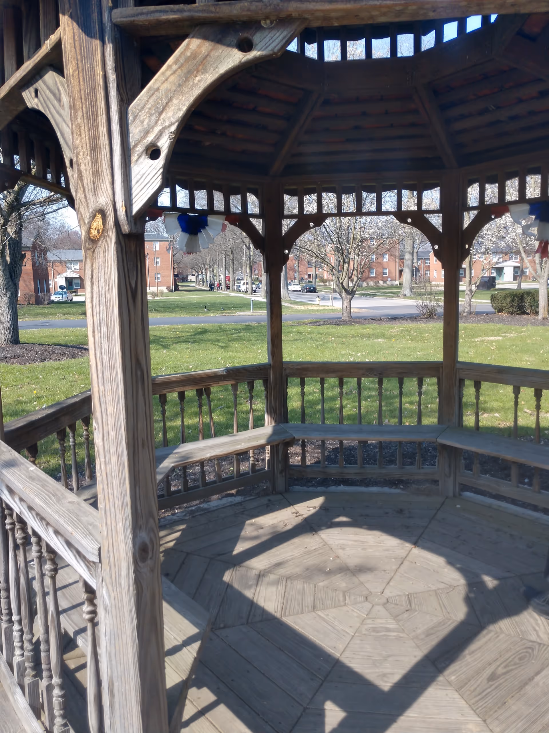 View from inside a wooden gazebo with built-in benches, located in a grassy outdoor area with trees and brick buildings in the background under a clear sky.