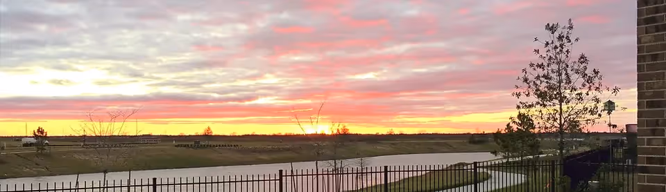 Sunset over a small lake with a metal fence in the foreground and a tree on the right.