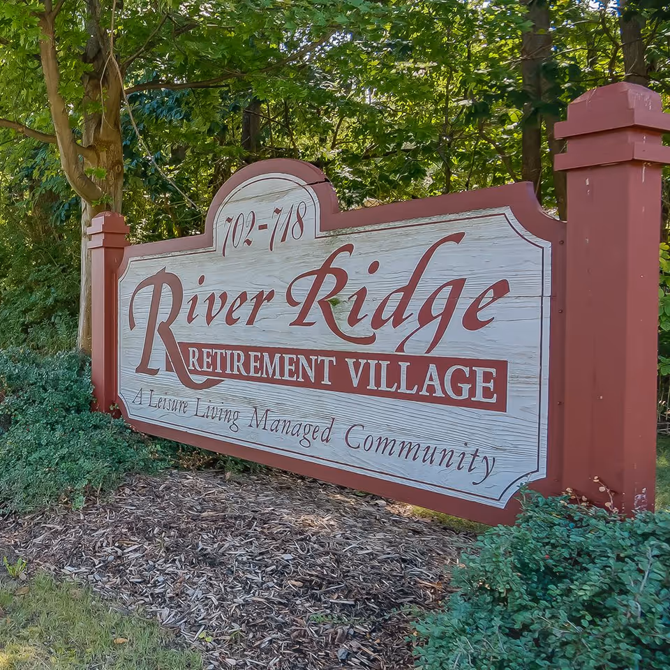 Wooden entrance sign reading "River Ridge Retirement Village" surrounded by trees and landscaping.