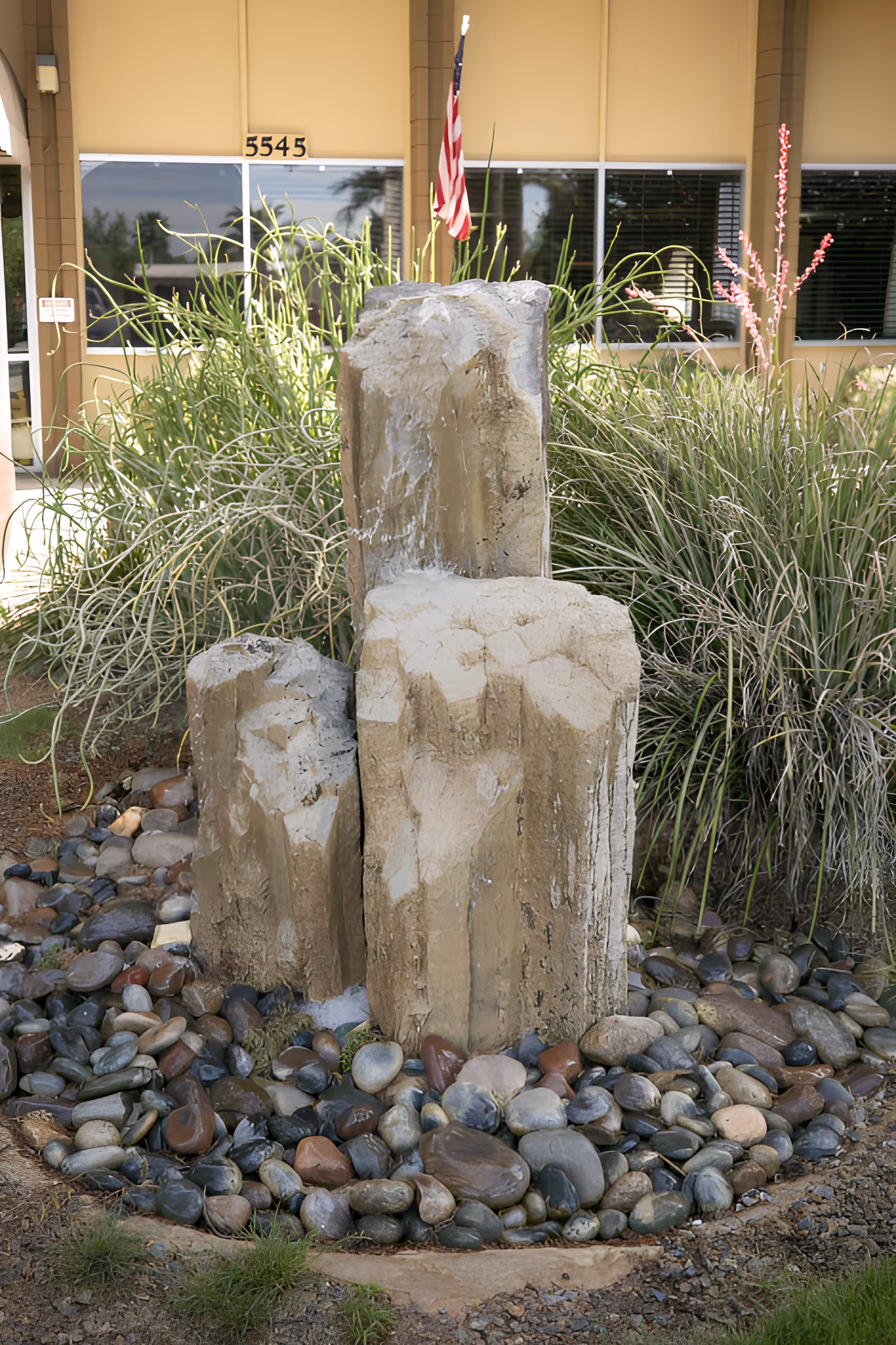 Outdoor water feature with three large vertical stone pillars surrounded by smooth river rocks and various green plants. An American flag is visible behind the stones, and a building with windows and the number 5545 is in the background.