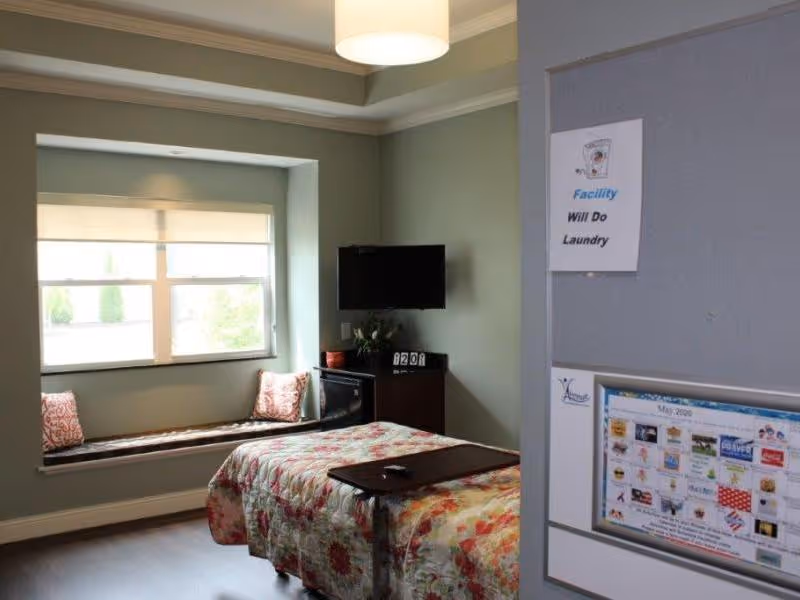 A senior-care bedroom with a floral-covered bed, window seat with cushions, wall-mounted TV, and a bulletin board on the door.