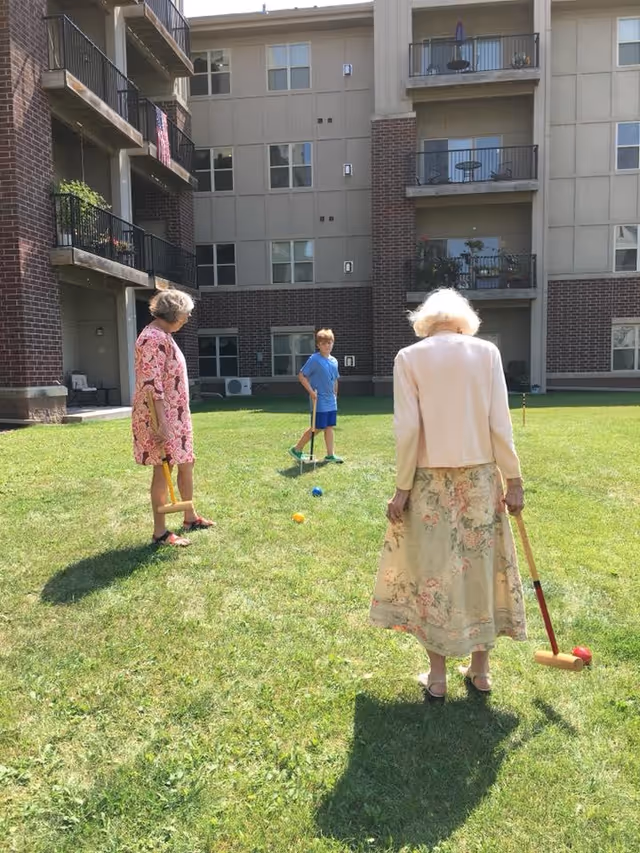 Two elderly women and a young boy playing croquet on a grassy lawn in the courtyard of a multi-story residential building with balconies and windows.