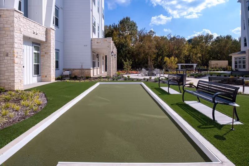 Outdoor bocce ball court with two black metal benches on artificial grass, surrounded by a landscaped garden area and multi-story building with stone and white siding exterior under a blue sky with some clouds.