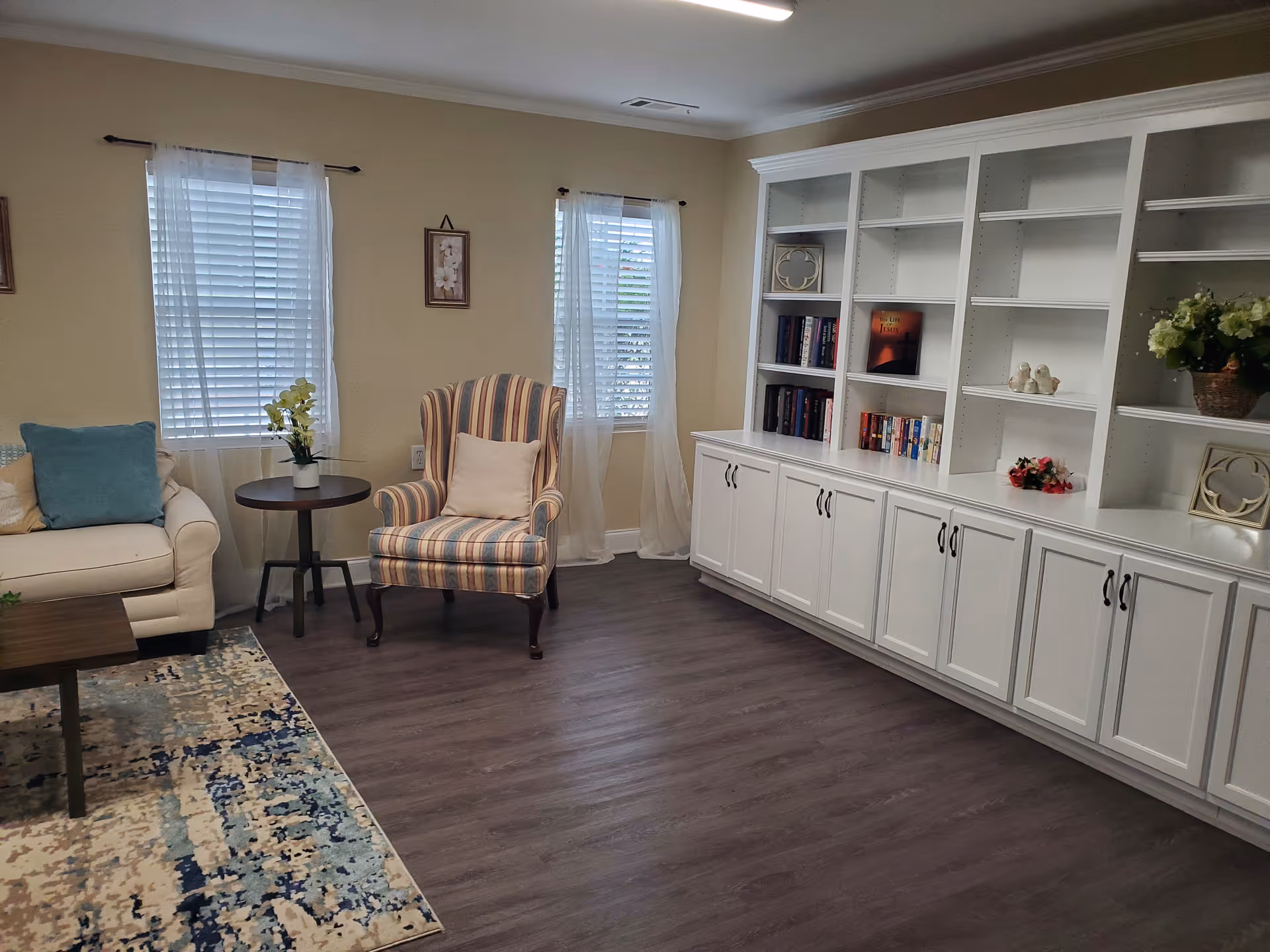 A cozy living room with a white couch adorned with blue and beige pillows, a round wooden side table with a small plant, a striped armchair with a beige pillow, two windows with white blinds and sheer curtains, a large white built-in bookshelf with books and decorative items, and a patterned area rug on a wood floor.