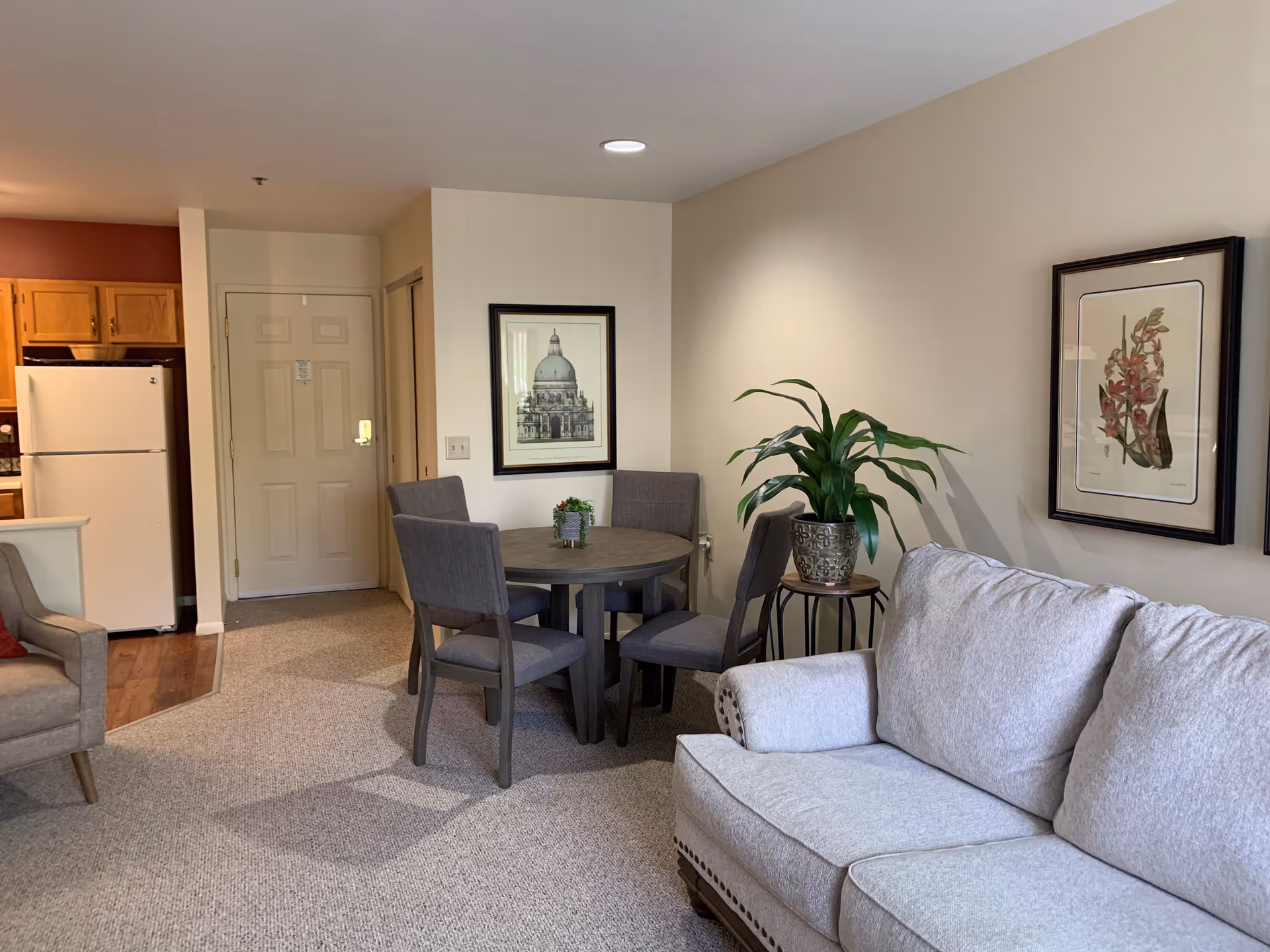 A cozy living area in a senior living facility featuring a light gray sofa, a round dining table with four gray upholstered chairs, a potted plant on a small side table, framed artwork on the walls, and a kitchen area with a white refrigerator in the background.