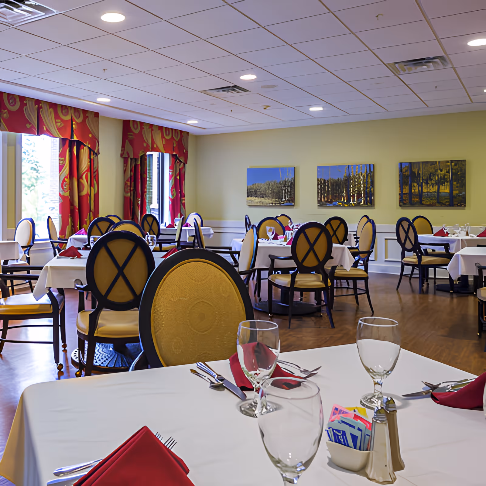 A dining room with multiple tables covered in white tablecloths, each set with red napkins, glassware, and silverware. The chairs have yellow upholstery with dark wooden frames. The room has large windows with red and gold curtains, light yellow walls adorned with landscape paintings, and a ceiling with recessed lighting.