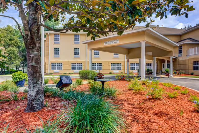 Exterior view of a senior living facility named Highland Place with a covered entrance, beige building walls, multiple windows, and landscaped garden beds with mulch, plants, and a large tree in the foreground.