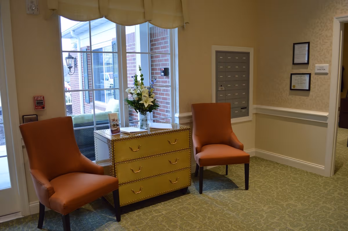 Two upholstered chairs flank a yellow chest topped with flowers in a well-lit lobby area with a window and wall mailboxes.
