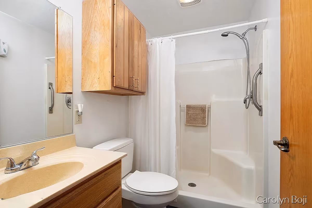 A clean bathroom featuring a beige countertop with a sink and faucet, a wooden cabinet above a white toilet, and a white shower area with a built-in seat, grab bars, and a white shower curtain. A beige towel hangs inside the shower.