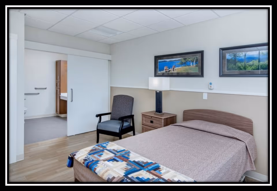 A senior care facility bedroom with a single bed covered in a beige quilt and a colorful patchwork blanket at the foot. Next to the bed is a wooden nightstand with a modern lamp. A cushioned armchair is placed beside the nightstand. The room has light-colored walls with two framed landscape pictures hanging above the bed. An open sliding door reveals an adjacent bathroom with grab bars and a wooden cabinet.