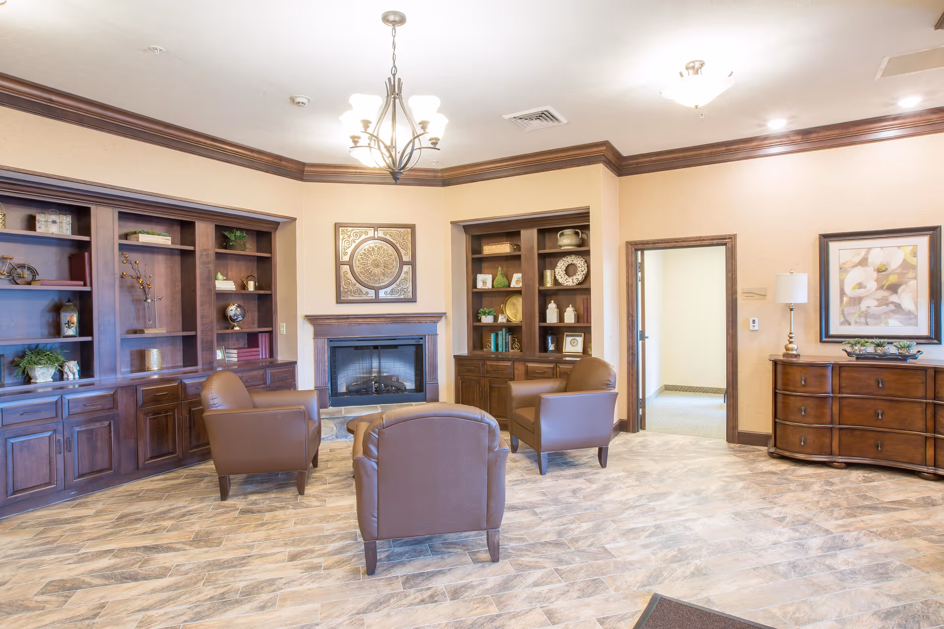 A cozy living room area with three brown leather armchairs arranged around a fireplace. The room features built-in wooden bookshelves filled with decorative items and books, a wooden dresser with a lamp and framed artwork above it, and a chandelier hanging from the ceiling. The floor has a light wood-patterned tile, and there is an open doorway leading to another room.
