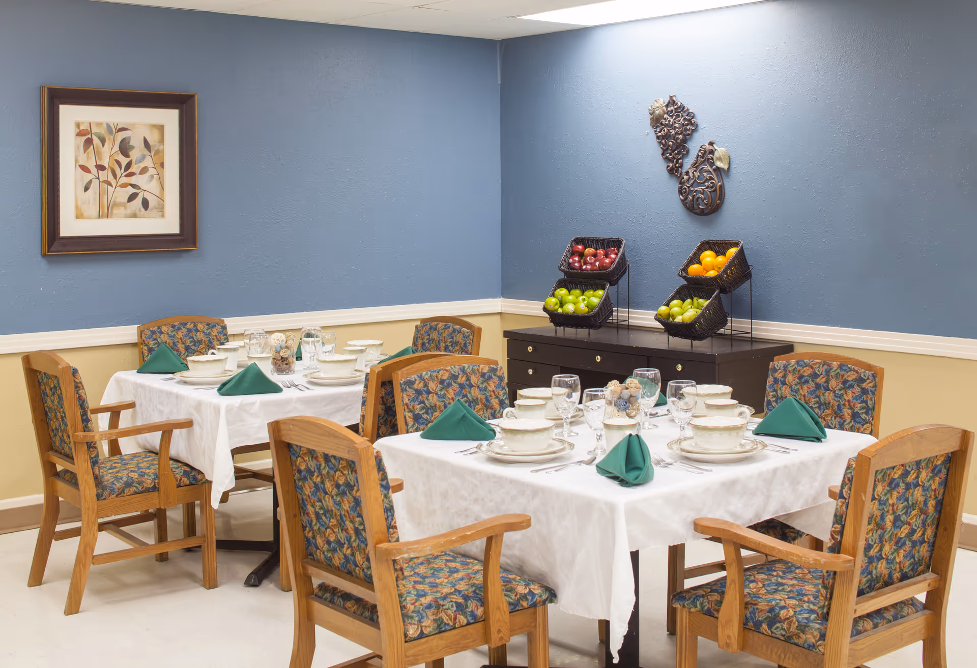 A dining room with two tables covered in white tablecloths, each set with bowls, plates, glasses, and green folded napkins. The chairs have wooden frames and floral-patterned cushions. On the blue wall, there is a framed artwork of leaves and a decorative metal wall hanging above a dark wooden sideboard holding baskets of apples and oranges.