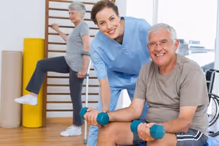 A smiling elderly man sitting and lifting dumbbells with the assistance of a female healthcare worker in blue scrubs, while an elderly woman exercises in the background in a rehabilitation or healthcare facility.