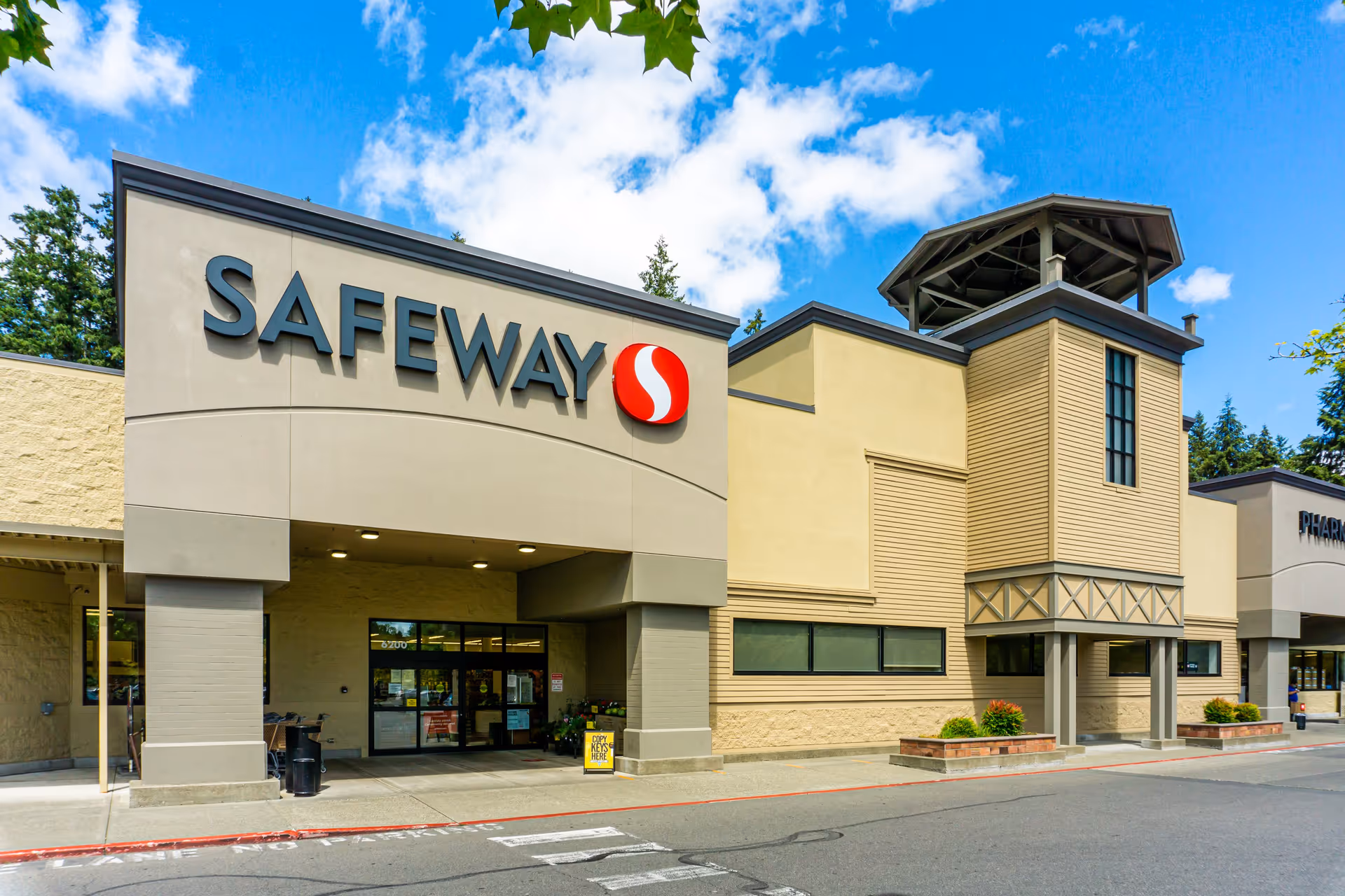 Exterior view of a Safeway grocery store with a large sign displaying the Safeway logo and name above the entrance. The building has beige and tan walls with a tower-like structure on the right side. The sky is blue with some clouds and there are trees visible in the background.