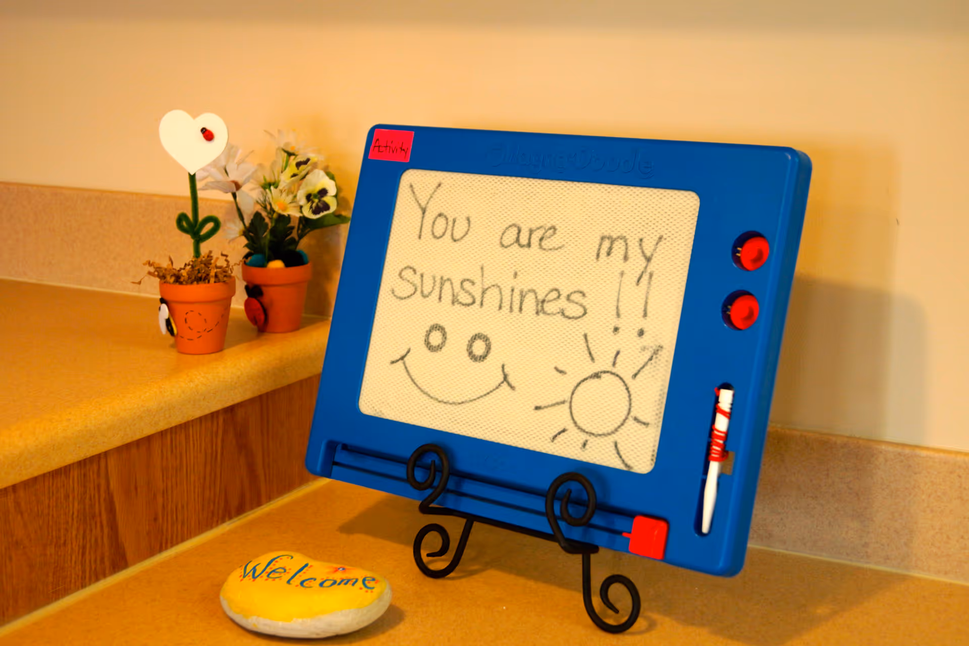 A blue Magna Doodle drawing board on a stand with the message 'You are my sunshines!!' written on it, accompanied by a smiley face and a sun drawing. The board is placed on a countertop next to two small potted plants and a painted rock with the word 'Welcome'.
