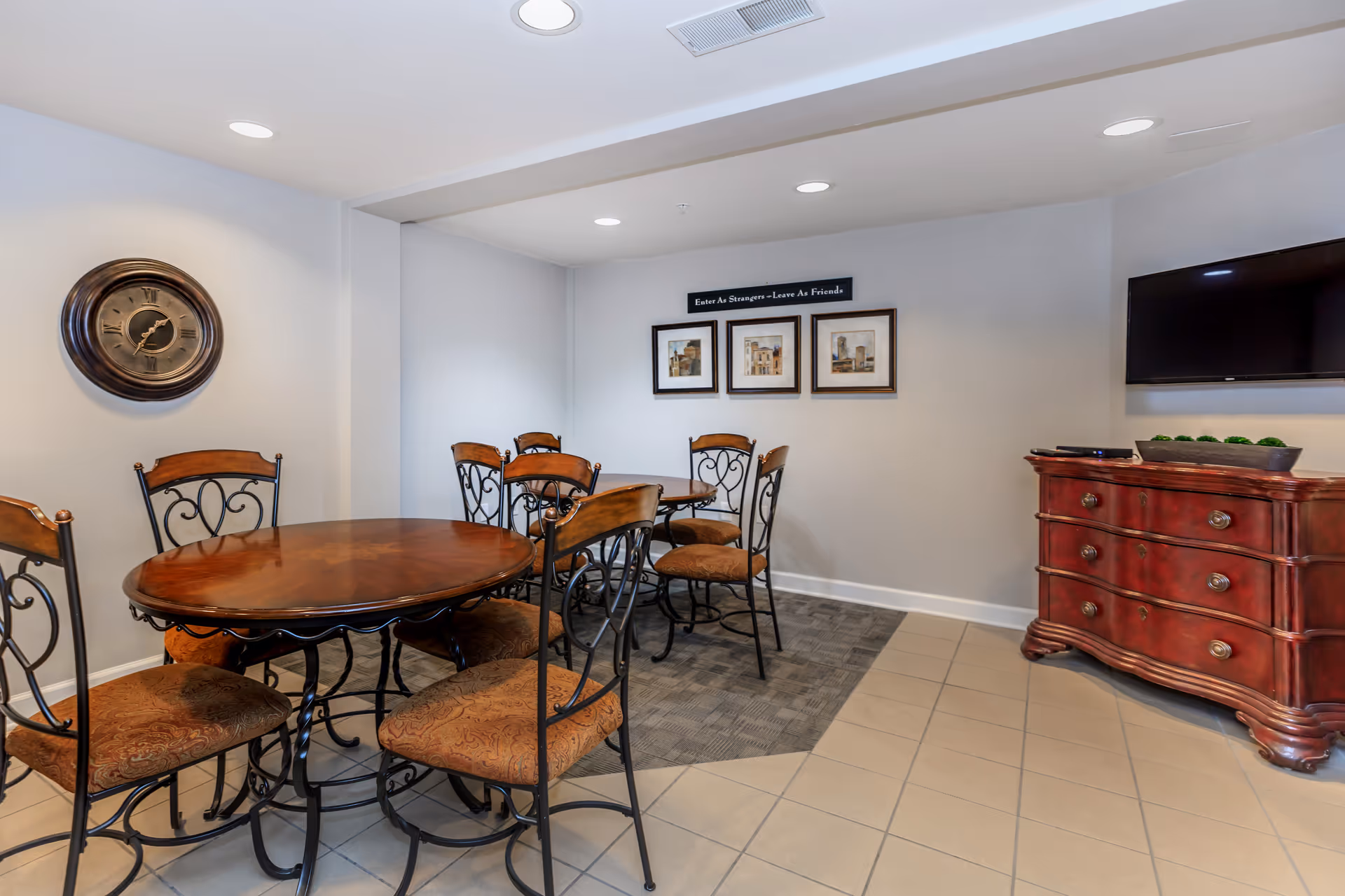 Small dining area with round wooden tables, wrought-iron chairs, a wall clock, framed artwork, and a flat-screen TV on a red dresser.