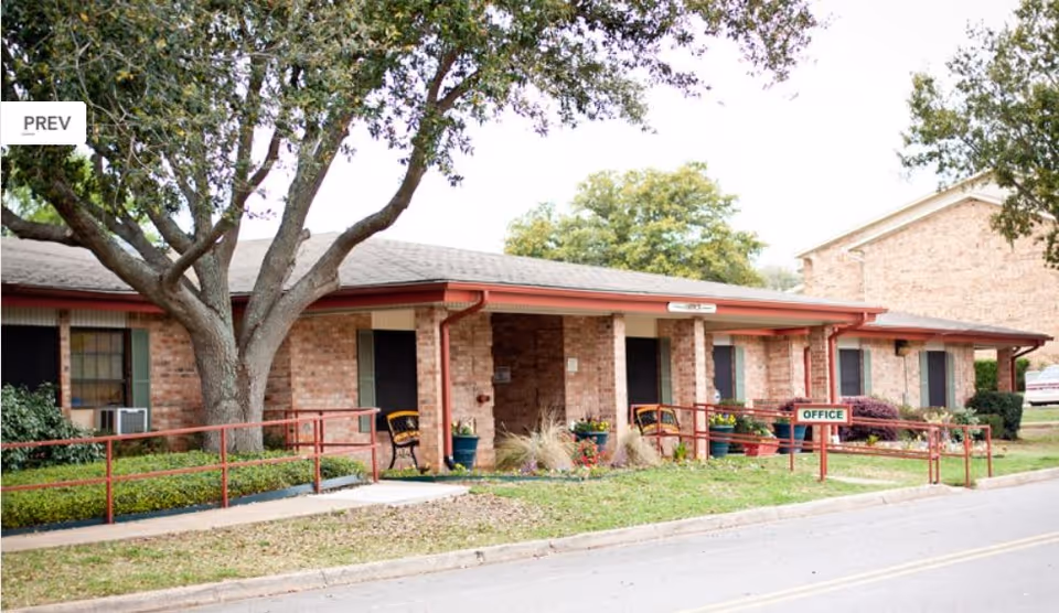 Single-story brick building front with a ramp, large tree, potted plants, and an 'Office' sign.