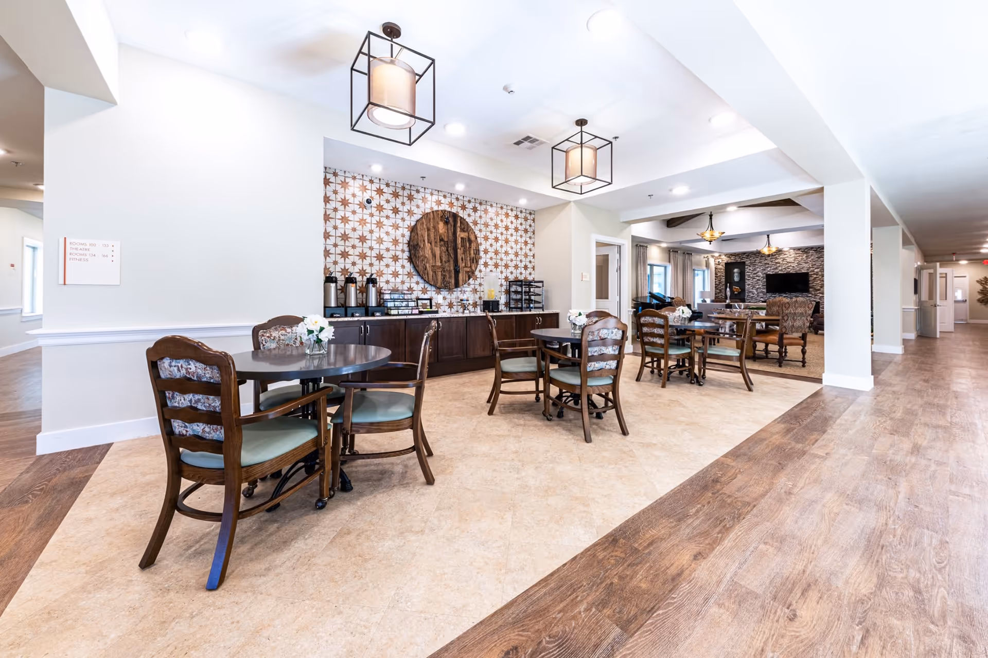 A bright and spacious common area in a senior living facility featuring several round tables with wooden chairs, each table decorated with a small vase of white flowers. The back wall has a decorative tile pattern with a large circular wooden art piece. There is a counter with coffee dispensers and other refreshments. The room has modern ceiling lights and a mix of tiled and wood flooring. In the background, there is a lounge area with a stone accent wall and a television.
