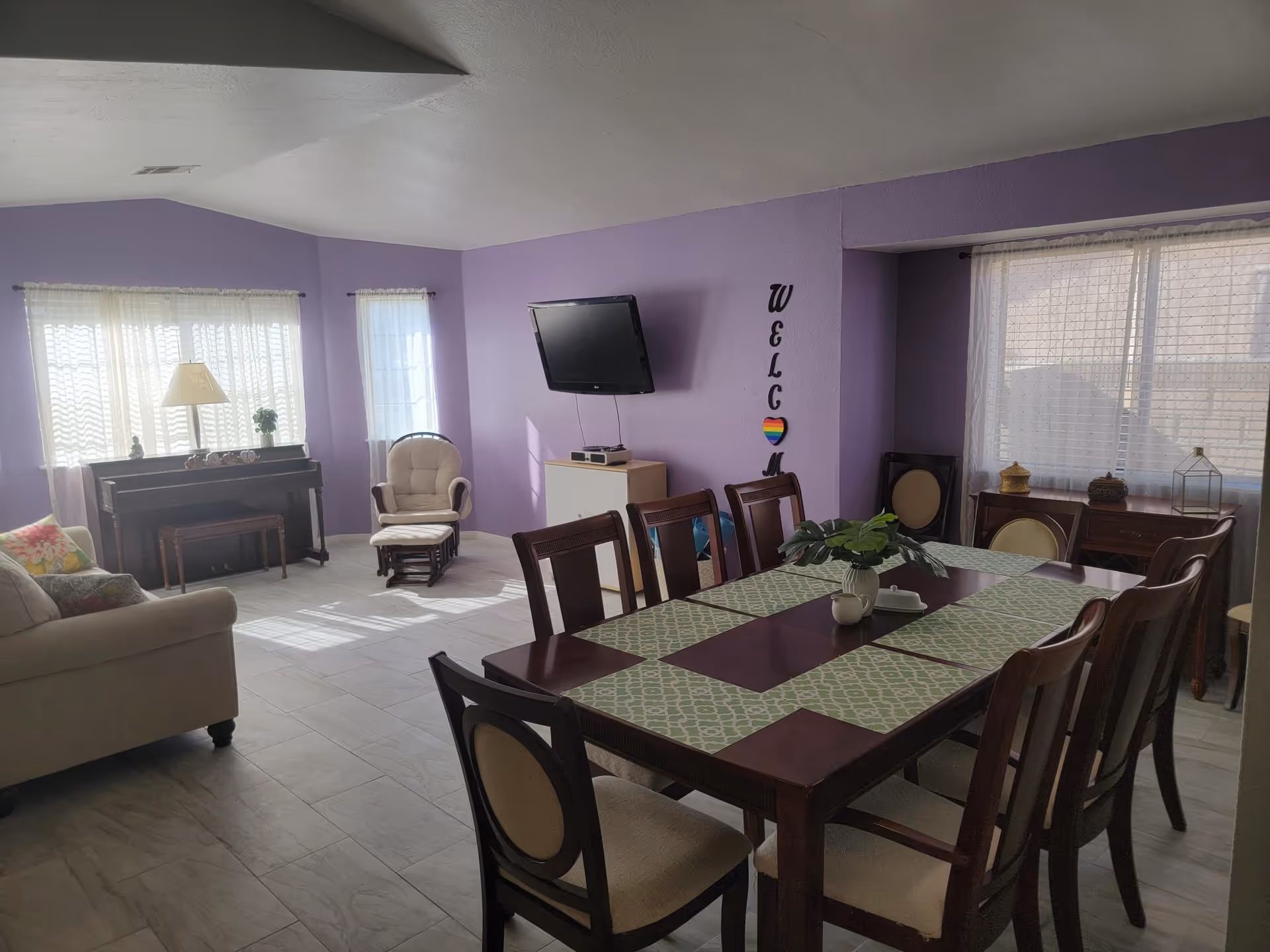 A bright living and dining area with lavender walls and large windows covered with sheer white curtains. The room features a wooden dining table with eight chairs and green patterned placemats, a beige sofa with colorful cushions, a rocking chair with an ottoman, a piano with a bench, and a wall-mounted flat-screen TV. The word 'WELCOME' is vertically displayed on the wall with a heart featuring rainbow colors.