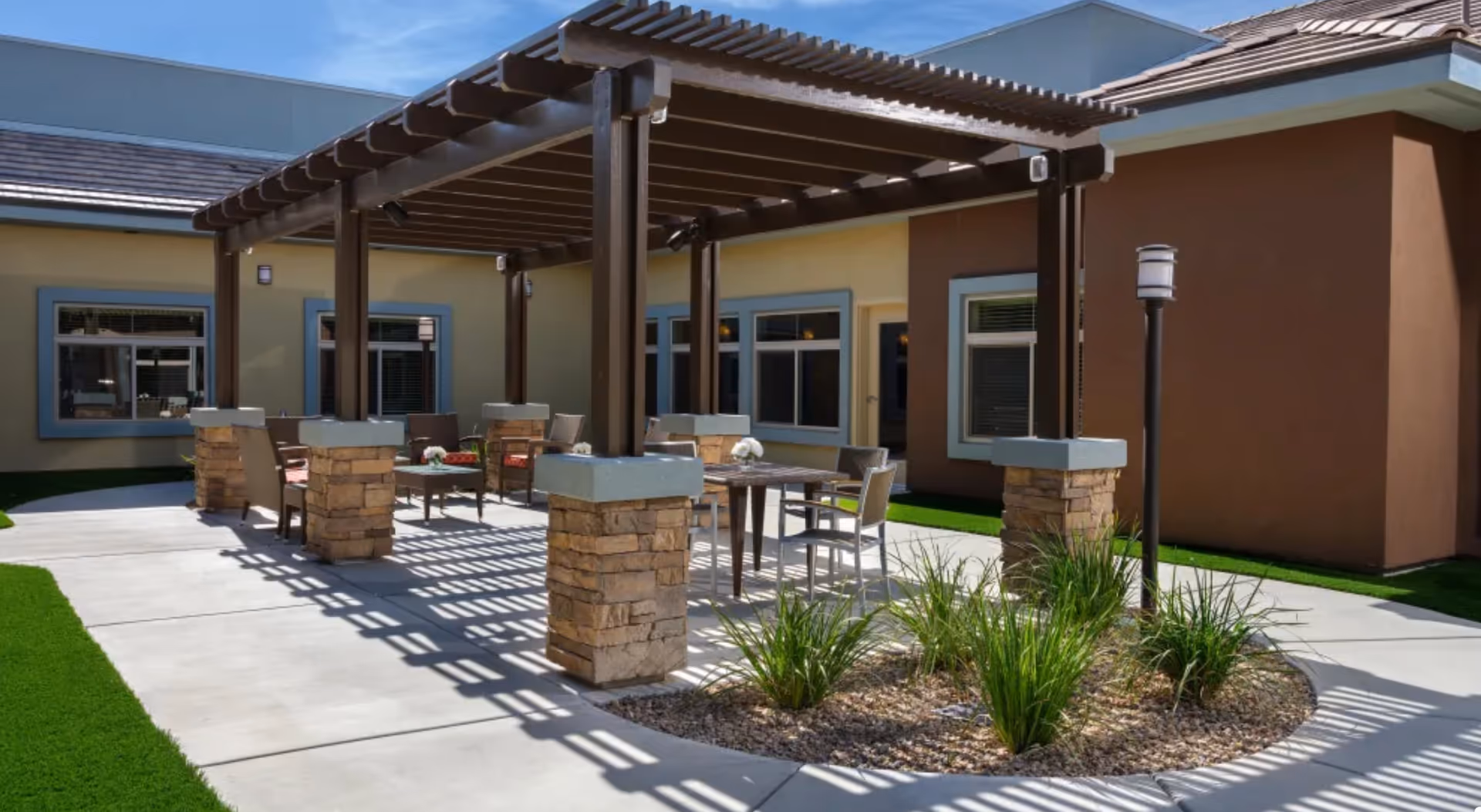 Outdoor patio area at Avenir Memory Care Westside featuring a pergola with stone pillars, several tables and chairs arranged underneath, surrounded by a concrete walkway and landscaped with green grass and plants.