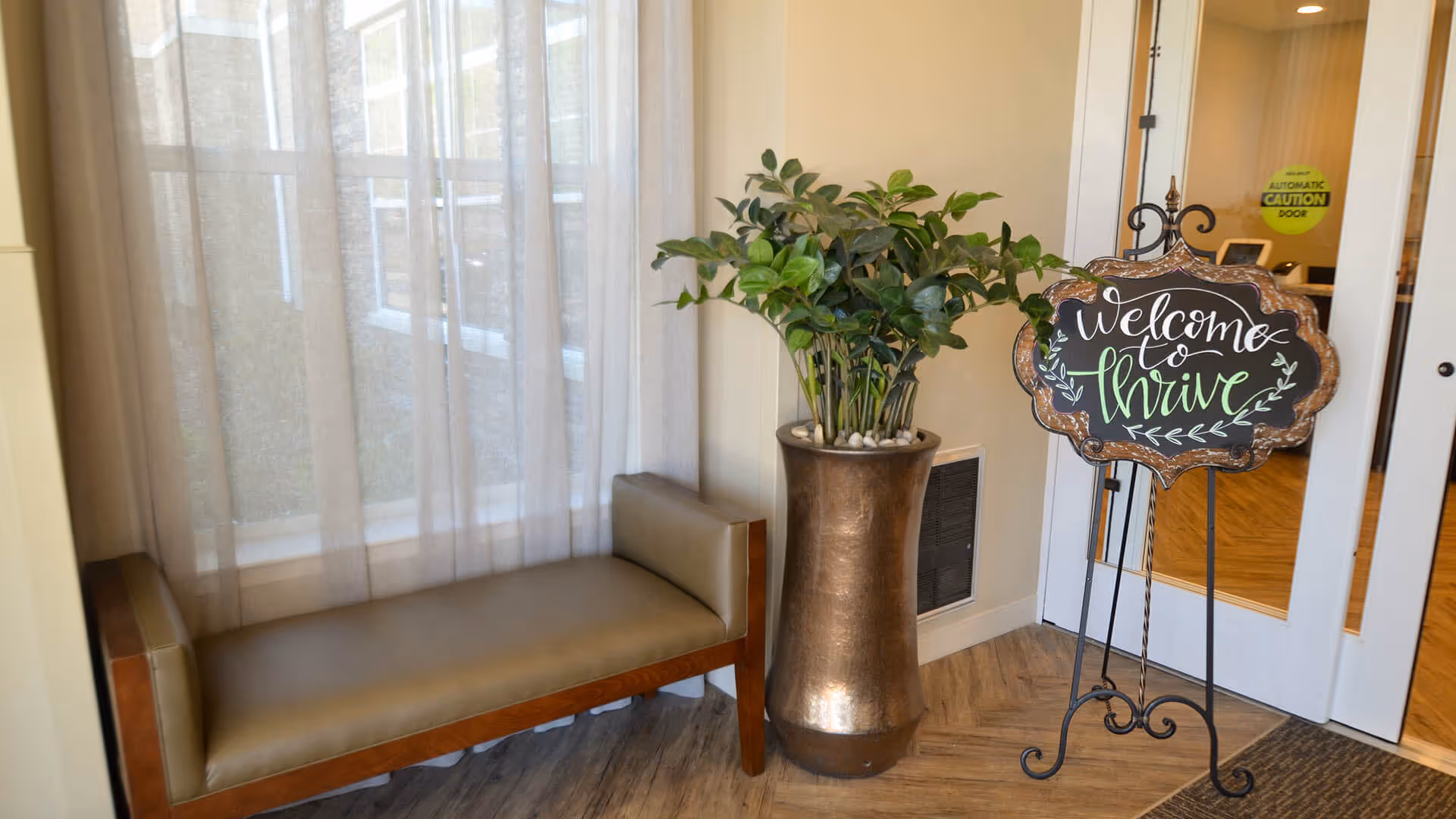 A cozy corner in a facility with a cushioned bench next to a window covered with sheer curtains, a tall bronze vase with a green leafy plant, and a decorative sign on a stand that reads 'Welcome to Thrive' near a glass door with an automatic caution sticker.