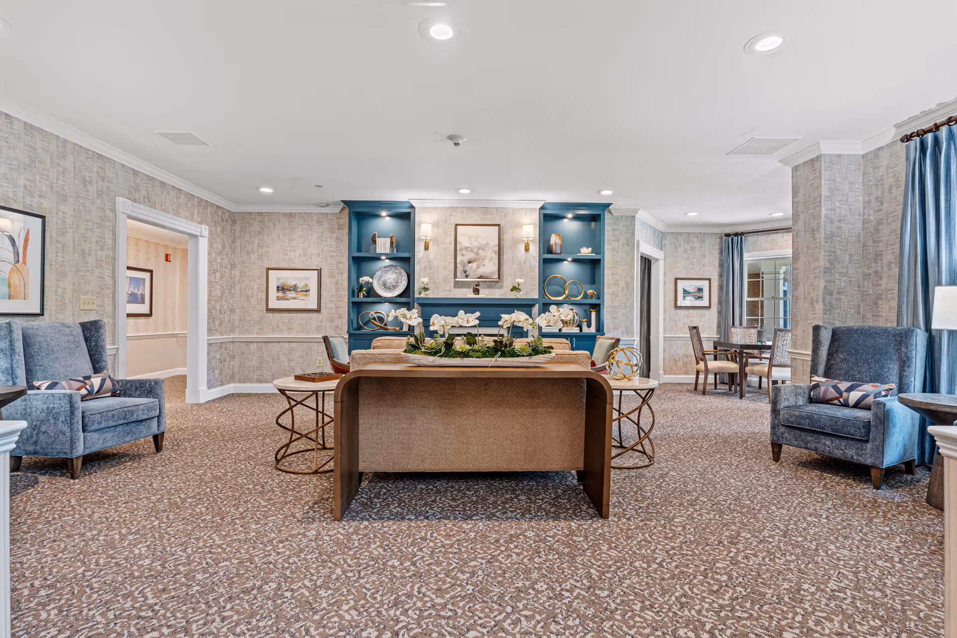 A spacious and well-lit living room area in a senior living facility with patterned carpet, two blue upholstered armchairs with decorative pillows, a beige sofa, and a wooden table with a floral centerpiece. The back wall features built-in blue shelves with decorative items and framed artwork above a fireplace. There is a small dining table with chairs in the background near windows with blue curtains.