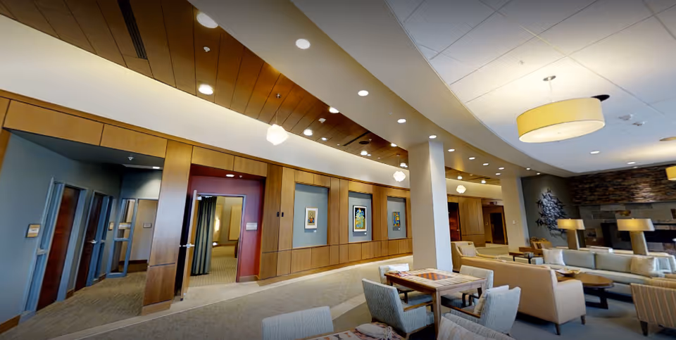 A spacious and well-lit common area in a senior living facility featuring comfortable seating with sofas and armchairs, a stone fireplace, modern ceiling lights, and wooden paneling on the walls. There are tables with chairs arranged for socializing or activities, and several doorways leading to other rooms.