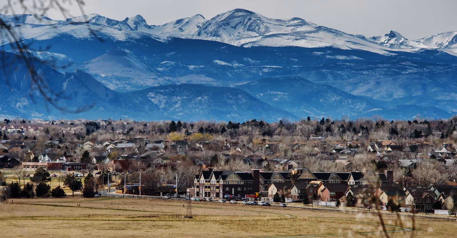 Distant view of a suburban neighborhood and a cluster of buildings in front of open fields with snow-capped mountains in the background.