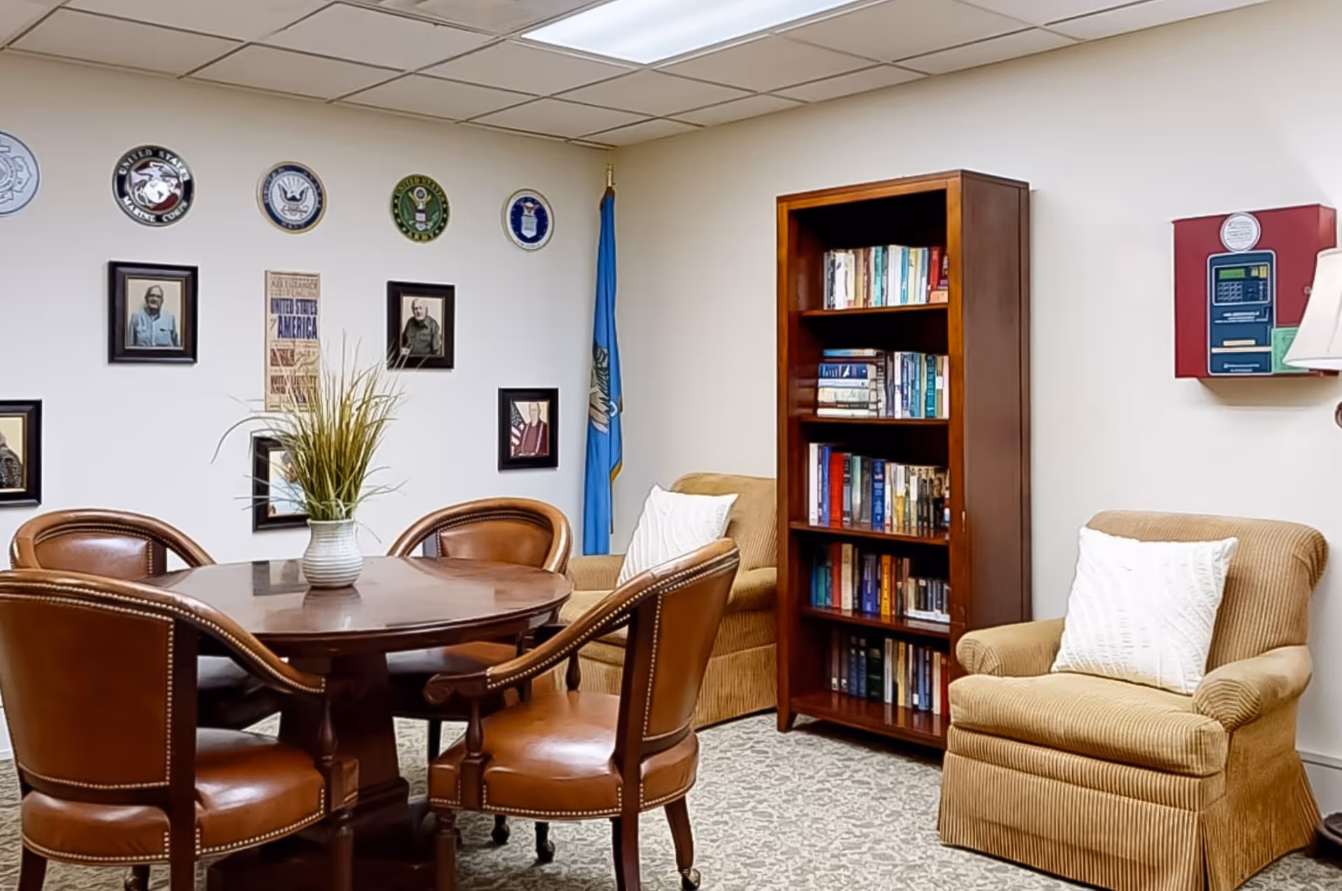 A cozy room with a round wooden table surrounded by four brown leather chairs. There are two beige armchairs with white pillows, a tall wooden bookshelf filled with books, and a blue flag in the corner. The wall is decorated with framed pictures and military insignias.