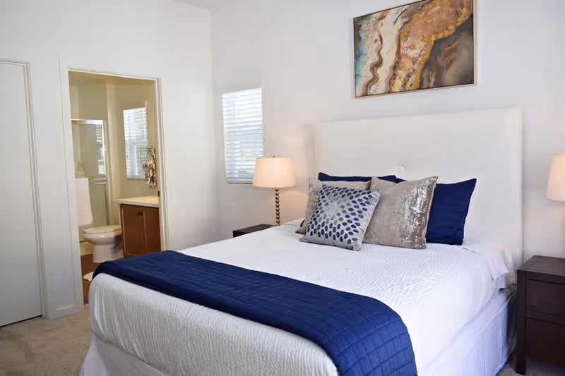 A bedroom with a neatly made bed featuring white bedding and a navy blue throw blanket. The bed has multiple decorative pillows in silver and navy blue. There are two bedside tables with lamps on each side of the bed. A piece of abstract art hangs above the headboard. To the left, an open door reveals a bathroom with a toilet, sink, and window with blinds.