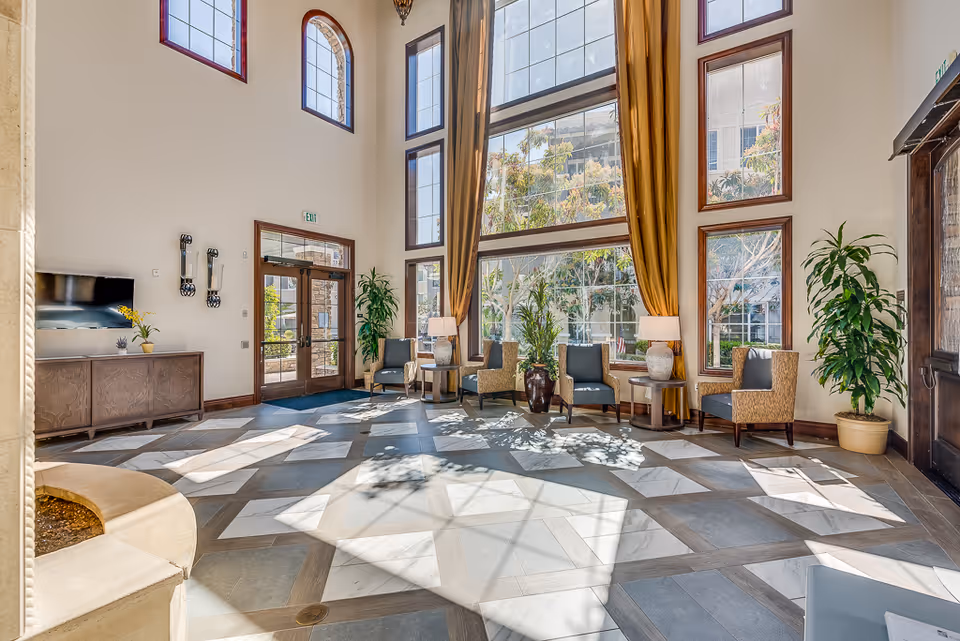 Sunlit senior living facility lobby with tall windows, seating areas, plants, and a patterned tile floor.