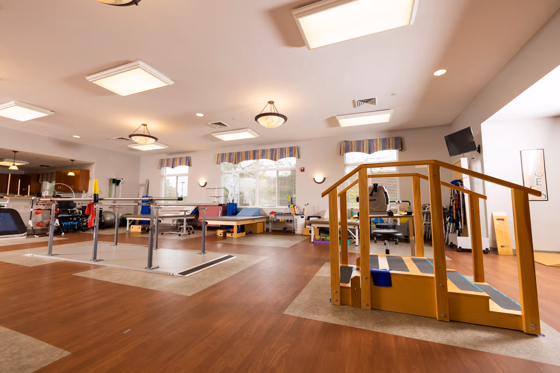 Well-lit interior therapy room with parallel bars, wooden practice stairs, exercise equipment and large windows.