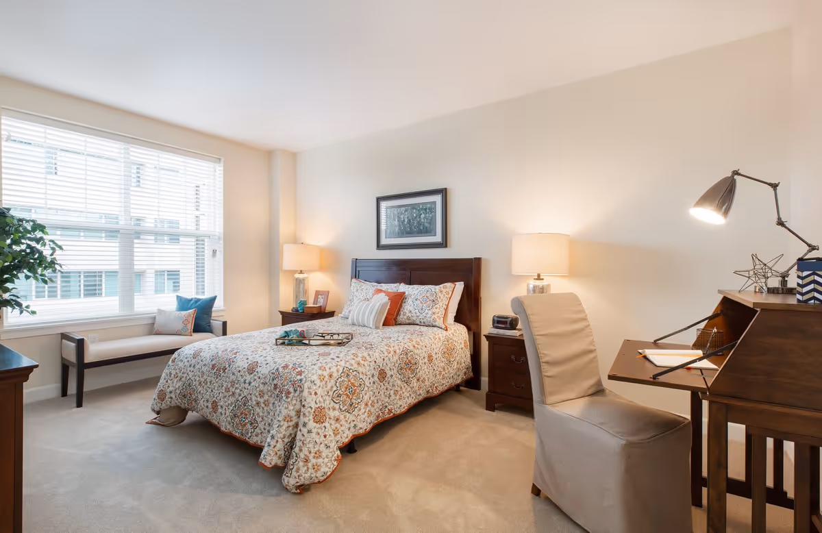 A bright and cozy bedroom with a large window covered by white blinds. The room features a bed with a patterned quilt and multiple pillows, two bedside tables each with a lamp, a cushioned bench by the window, a wooden desk with a modern lamp, and a beige upholstered chair. The walls are painted light beige and the carpet is light-colored.