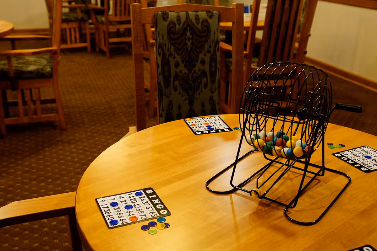 A round wooden table with a bingo cage containing colorful balls and several bingo cards with chips placed on the table. The background shows wooden chairs and a carpeted floor in a cozy indoor setting.