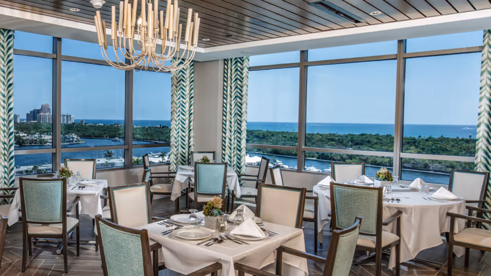 Bright dining room with set tables, chairs and a chandelier overlooking water and greenery through large windows.