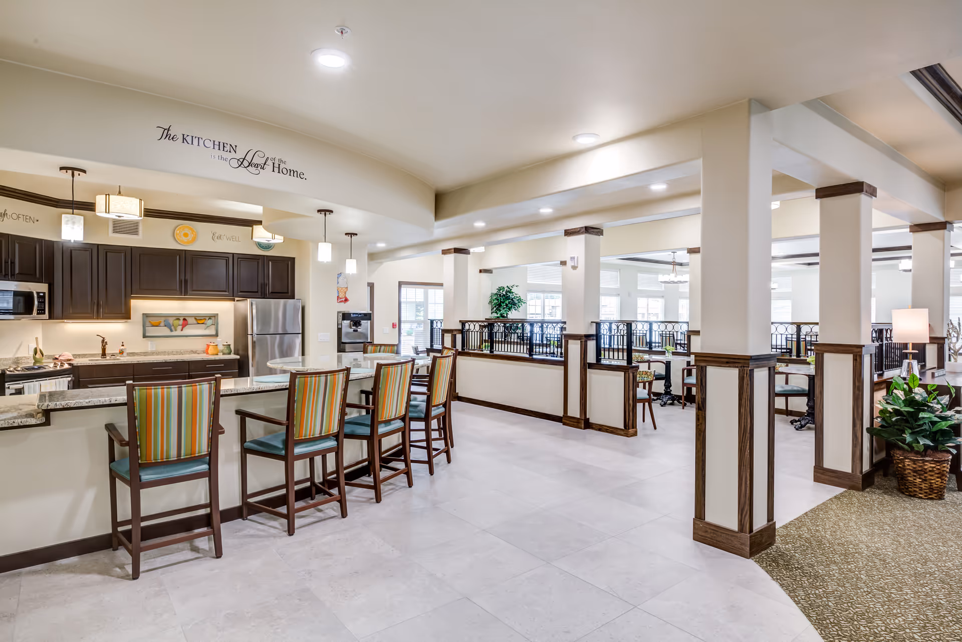 A bright and spacious kitchen area in a retirement living facility with a granite countertop bar and four striped cushioned chairs. The kitchen features dark wood cabinets, stainless steel appliances including a refrigerator and microwave, and pendant lights hanging from the ceiling. Adjacent to the kitchen is a dining area with tables and chairs, separated by decorative railings and columns. The walls have decorative text, including a quote above the kitchen that reads 'The kitchen is the heart of the home.'