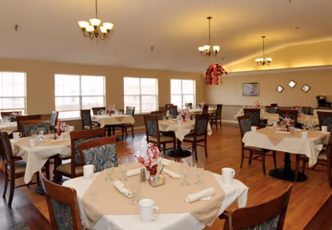 A spacious dining room with multiple tables covered in beige tablecloths and white napkins, each set with cups, glasses, and small floral centerpieces. The room has wooden floors, several windows letting in natural light, and ceiling lights with chandeliers. Chairs with patterned upholstery surround the tables.