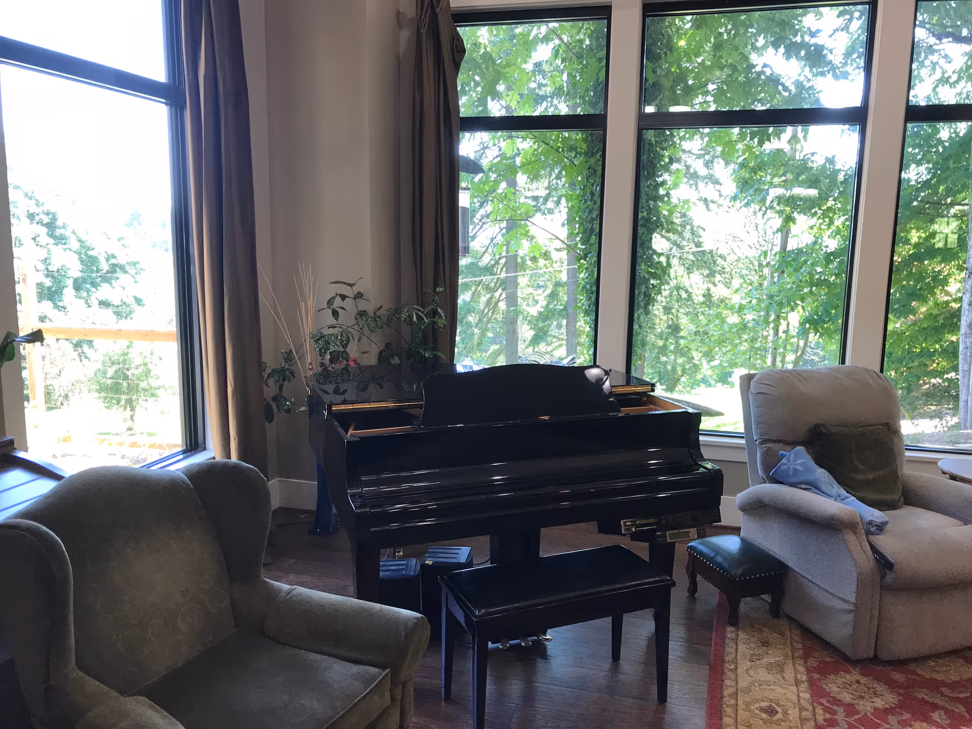Sunlit living room with a black grand piano, two upholstered armchairs, and large windows looking out to trees.