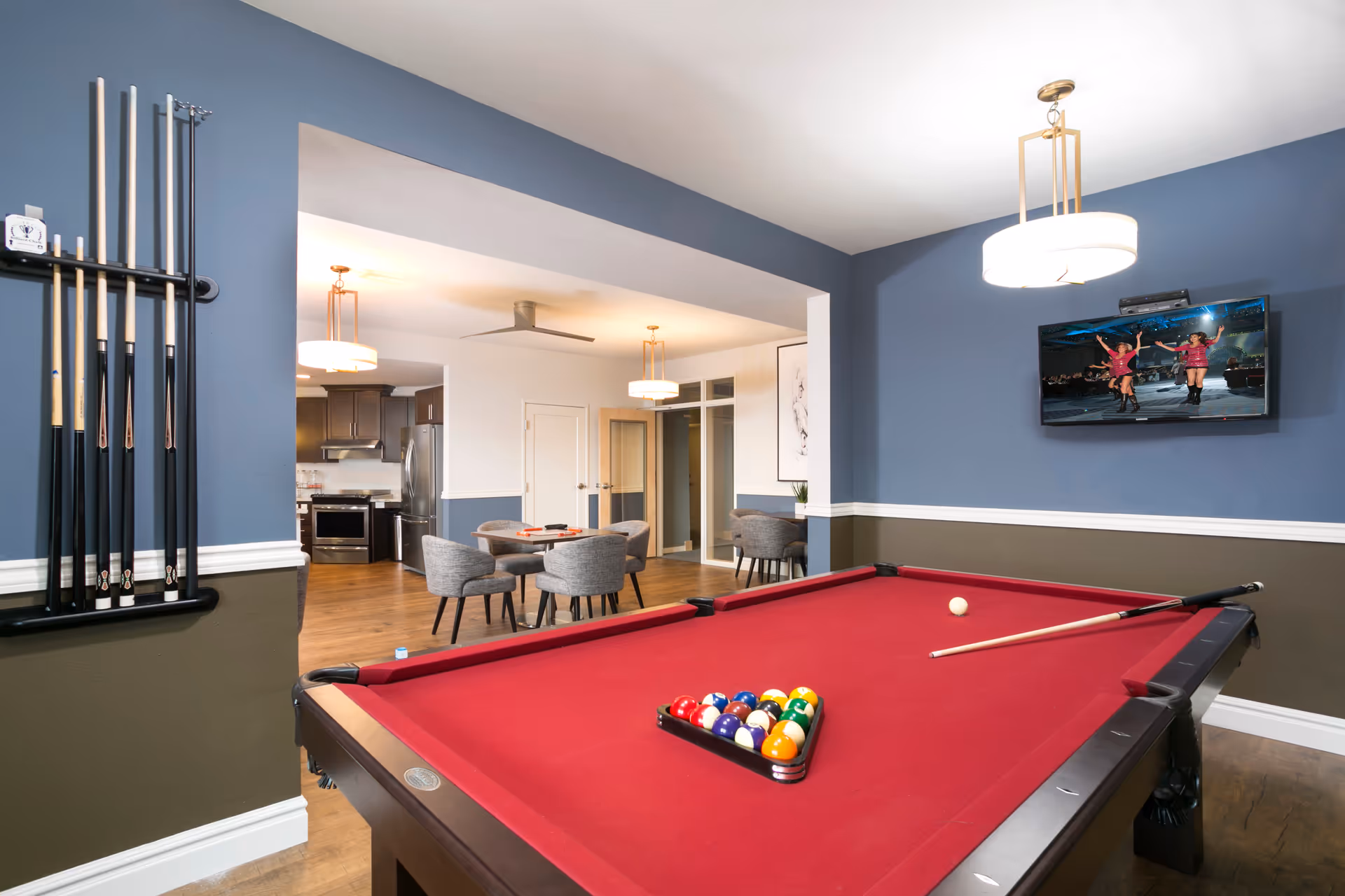 A recreational room with a red pool table in the foreground, pool cues mounted on the left wall, and a flat-screen TV mounted on the right blue wall showing a dance performance. In the background, there is a dining area with a round table and four gray upholstered chairs, and a kitchen with stainless steel appliances.