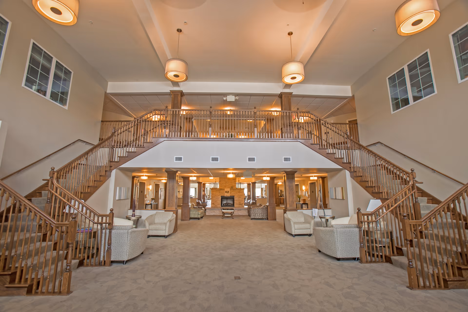 Spacious interior of a senior living facility featuring a large open area with carpeted floors, multiple beige armchairs arranged in seating groups, a central fireplace, and a grand double staircase with wooden railings leading to an upper balcony. The ceiling has modern pendant lights, and there are windows on the upper walls allowing natural light.