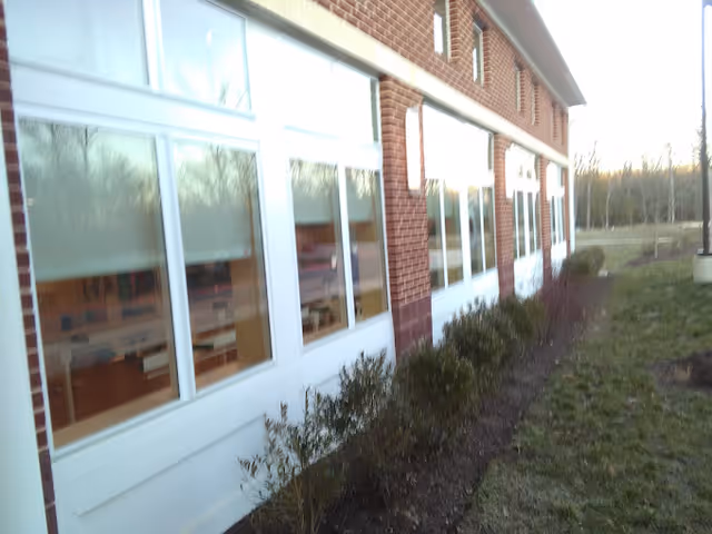 Exterior view of a brick building with large windows and small bushes planted along the side. The building is adjacent to a grassy area and trees are visible in the background.