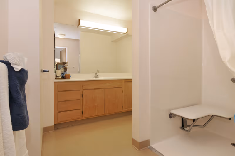 A bathroom with a wooden sink vanity and mirror, and a walk-in shower with a fold-down bench and towel on the wall.