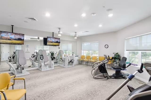 A bright and spacious fitness room with exercise equipment including stationary bikes and weight machines. The room has large windows with white blinds, a wall clock, yellow chairs lined up against the wall, and two flat-screen TVs mounted on the mirrored wall.