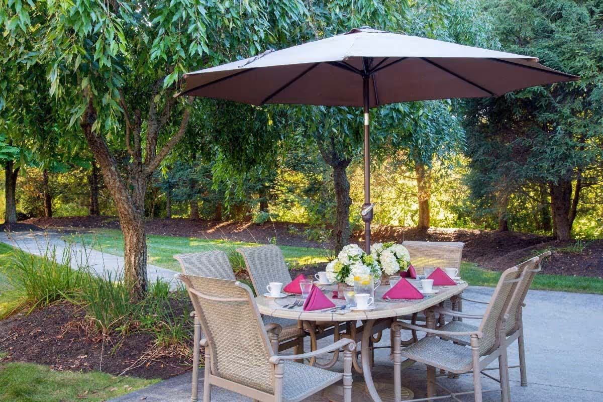 Outdoor patio dining table with umbrella and chairs set with pink napkins and a floral centerpiece beneath trees.