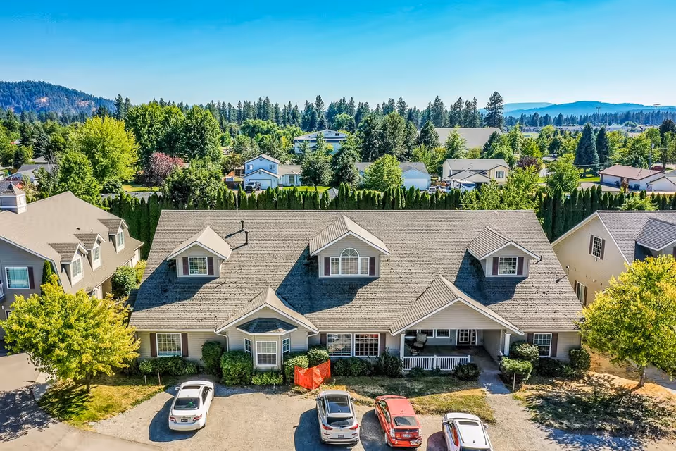 Aerial view of a senior living facility building with a gray roof and beige exterior, surrounded by trees and greenery. There are several cars parked in front of the building, and neighboring houses and trees are visible in the background under a clear blue sky.